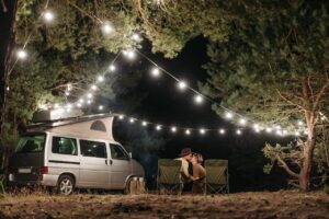 Couple sitting by a campervan enjoying a romantic night under string lights in a forest.